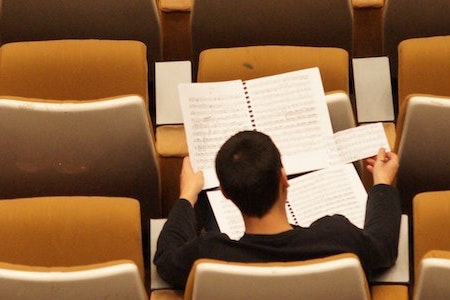 Image of rows of seats in an auditorium, seen from above. One person, studying a musical score and wearing a black long-sleeved shirt, is visible sitting in one of the seats.